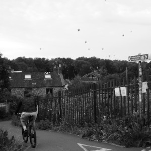 A cyclist next to a fenced off allotment goes downhill. Hot air ballons are peppered throughout the sky.
