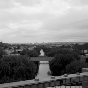 A canal with leafy trees around. Bridges cross the canal. City buildings are visible in a summer haze.