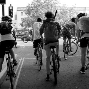 A group of cyclists are stopped by a traffic light. Most are wearing shorts and t-shirts, most have a foot on the ground.