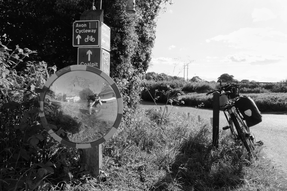 A black and white self-portrait. I am a white woman with a helmet on and a camera in front of my face crouched in front a road mirror beneath cycle path signs. My bicycle rests on a post. Beyond a lane with leafy bushes can be seen.