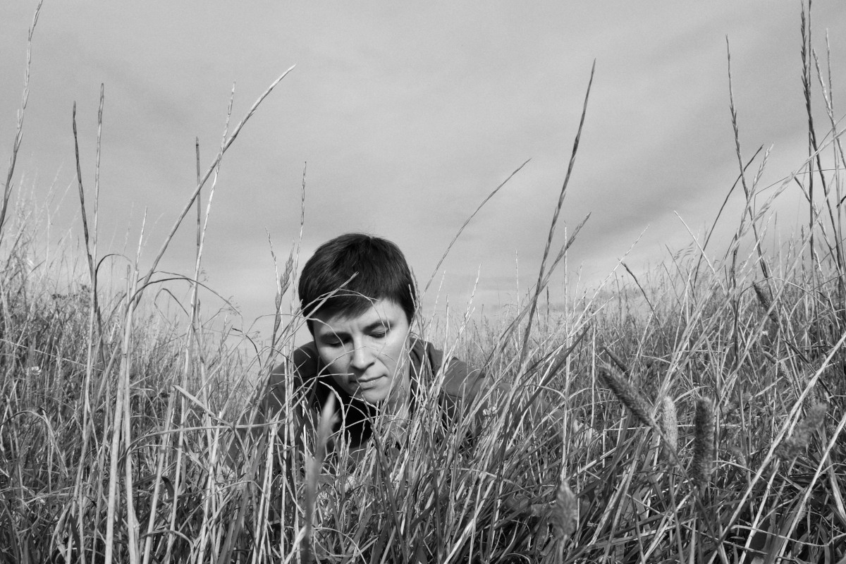 A black and white self-portrait. I am a white woman with short dark hair, lying in tall grass. Only my shoulders and head are visible through a curtain of grass. Above, the sky is full of clouds.