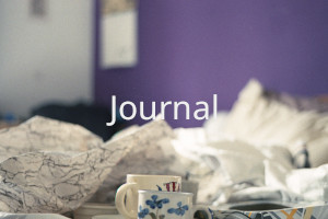 An unmade bed with wrapping paper and mugs on it. Over the image the word 'Journal' is written in uppercase white letters.