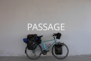 A baby blue bicycle loaded with black panniers rests against a greyish wall. Over the image the word 'Passage' is written in uppercase white letters.