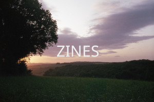 A green field bordered by rounded tree top and a sunset in the distance on the left hand side. Over the image the word 'Zines' is written in uppercase white letters.
