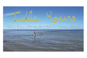 A view of the sea with two children on the water. One if lying down, the other standing. In the sky are the handwritten large words 'Idle Hours'. Below in a smaller script is 'Stills from the holiday movie I never shot'.