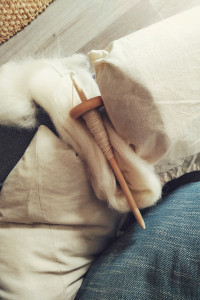 A vertical colour photograph of a wooden drop spindle with white spun wool on the shaft. It rests on a bit of white wool fibre but a white linen bag. The whole rests on a grey sofa with a white pillow and a turquoise pillow. The floor beyond the sofa is a light grey brown. In the top far left corner a bit of a jute mat is visible.