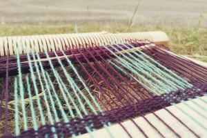 A horizontal colour photograph of a small wooden loom threaded with purple and turquoise yarn. The weaving is only a handful of row deep.