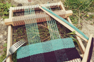 A horizontal colour photograph of a small wooden loom threaded with purple and turquoise yarn. The weaving alternates a thick row of purple and a thick row of turquoise. The loom rests on pavement slabs with a lot of grass and greenery pocking through the cracks.