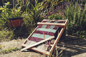 A horizontal colour photograph of a small wooden loom resting against a small grey flower pot. All around is greenery. There is a baby oak in a pot, lavender, weeds, rosemary, etc.