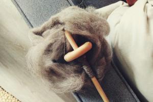 A horizontal colour photograph of a wooden spindle resting on a pile of grey wool. There is a small cop on the shaft. The wool and spindle rest on the armrest of a grey sofa. There is a bit of white pillow visible to the left and a light coloured wooden floor on the left.