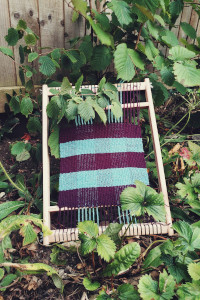A vertical colour photograph of a small wooden frame loom with a checked woven fabric in purple and turquoise. The loom rests against a couple of hazel trees by a wooden fence.