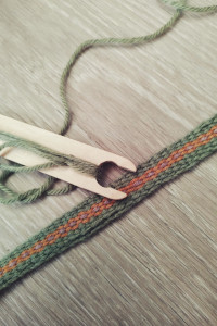 A vertical colour photograph of a woven band in green, orange, and mauve. The band is on a light coloured floor. Part of it is not woven well, another part is woven better. A wooden shuttle with green wool on it rests at the side of the band.