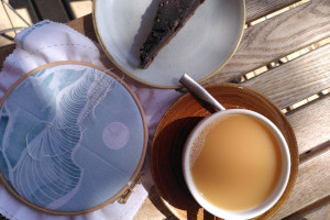 A horizontal photograph of an embroidery hoop by a chocolate and raspberry torte and a cup of earl grey tea on a wooden outdoors table.