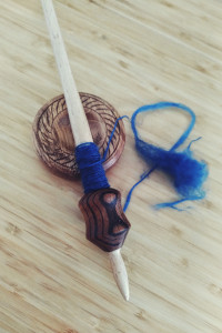 A vertical colour of a wooden supported spindle resting across its spinning bowl. There is some blue yarn spin on it. A little bit of blue unspun yarn lies next to it. The thread connecting the two is broken.