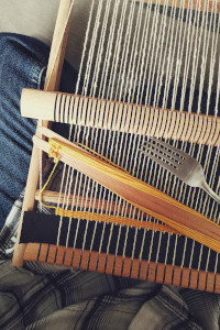 A vertical colour photograph of a frame loom resting on my crossed legs. I am wearing a pair of blu jeans and checkered black and white shirt. On the loom is the start of a weave using yellow cotton.