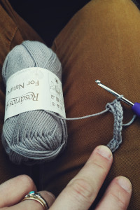 A vertical colour photograph of a crochet hook with a greyish ball of cotton yarn resting on my legs in a mustard jeans.