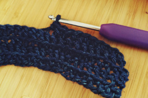 A horizontal colour photograph of a crochet hook on a desk. The hook is on a small crochet work using blue denim cotton. The crochet work is a small rectangle of single crochet.