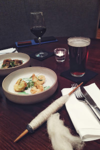 A vertical colour photograph of a pub table with a couple of round plates with nibbles in them. There is a pint of bitter and a small glass of red wine too. In front of it all is a wooden spindle with a small metal whorl and white wool spun on it.
