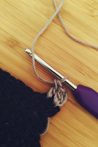 A vertical colour photograph of a the corner of a blue denim crochet work. A light purple border is starting to be crocheted on the denim. The crochet hook is attached to the yarn.
  