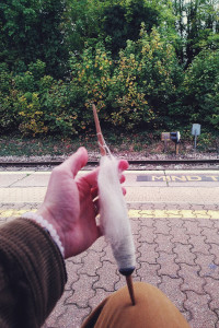 A vertical colour photograph of my hand holding a wooden spindle with white wool on it. The spindle tip is on my trousers (mustardy jeans). Behind is a small train station with rails below the platform edge and on the other side green bushy trees with fading leaves.
