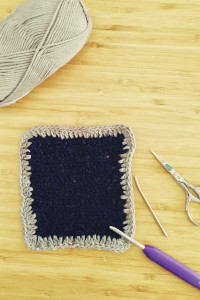 A vertical colour photograph of a crochet square. It is dark navy blue with a greyish lavender border. It's all wonky. Around it are a crochet hook, a pair of small scissors, a darning needle, and a ball of cotton used for the border.