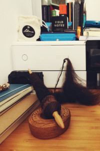 A vertical colour photograph of a wooden spindle full of dark brown spin wool. A bunch of unspin wool rests at the side. The spindle tip is resting in a small wooden bowl. At the other end is a white printer. Around the spindle on the desk are some notebooks, pens, and a camera. On the printer image more notebooks, a felt glass case, and random bits and bobs.