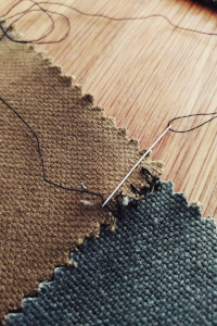 A vertical colour of a green brown piece of fabric being sewn with a needle and black thread to a blue grey piece of fabric.
