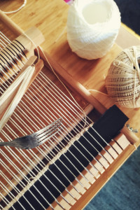 A vertical colour photograph of a corner or a flat wooden weaving loom wrapped with white thread. At the bottom, the weft has started in a fine creamy thread. On the wrap rest a shuttle loaded with thread and a fork. Next to the loom rests two balls of thread, one used for the wrap, the other used for the weft.