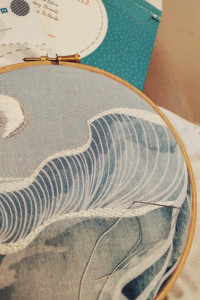 A vertical colour photograph of a pre-printed embroidery project. The fabric in the wooden hoop is shaded of soft blues with white and silver embroidery threads that create a moon and stylised waves. The stitching is not very neat.or precise.