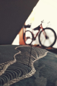 A vertical colour photograph of an ongoing embroidery project in white threads over a blue background. The motif is a stylised wave. In the background is a blurred bicycle.