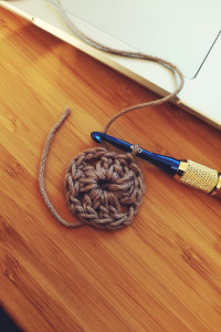 A vertical colour photograph of a blue crochet on a wooden handle. On the crochet is a lavender cotton yarn with the beginning of a granny square. The whole rests on a desk. Next to them, a laptop is visible but only part of the trackpad.