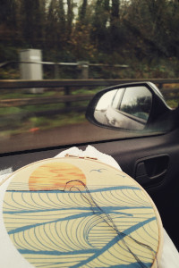 A vertical colour photograph of an embroidery loop on a car. The embroidery is a stylised sunset printed on cotton to guide the stitching. The hoop rests by the passenger window. Outside, vegetation and some barriers are blurred from the speed of the car.