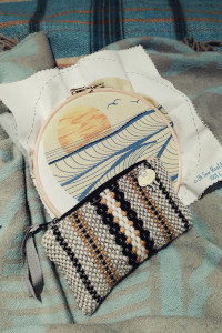 A vertical colour photograph of a pre-printed embroidery work resting on a bed. The embroidery is a stylised sunset over the sea. Some embroidery has started but the work is mostly empty of thread. A woven pouch rests in front of the hoop.