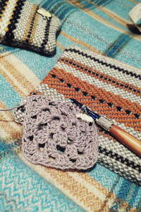 A vertical colour photograph of a lavender granny square with a crochet hook still attached to it. By the crochet hook is a stitch marker in the shape of a sheep. The work rests on a cream, orange, and black woven purse. A other smaller woven purse in yellow, black, and cream is visible at the top left of the image. The image is set on a background of a blue and yellow checkered blanket.