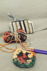 A vertical colour photograph of a colourful small granny square on a grey sofa. A crochet hook is still attached to the square. Behind is the yarn used. It's a colour changing yarn. Behind that is a woven yellow, cream, and black small pouch with a sheep pin in it.