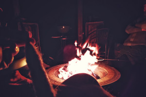 A horizontal colour photograph of a campfire. I am holding a spindle thick with wool in front of it. Above the flames someone is holding a metal stick with marshmallows on it.