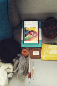 A vertical colour photograph of a spread of books, embroidery kit, yarn, combed wool, wooden spindle, handkerchief, and crochet hook.