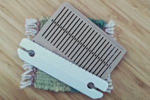 A square colour photograph of a 3D printed brown rigid heddle and white shuttle. They rest on a yellow and green coaster on a bamboo tabletop.