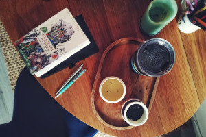 A horizontal colour photograph of a wooden table with a tray holding tea accoutrements, a couple of pens, a book (Medieval Bodies by Jack Hartnell), a black notebook mostly hidden by the book, and a light green candle.