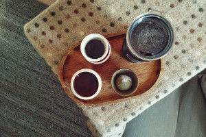 A horizontal top down colour photograph of a table covered with an oatmeal colour blanket. On top of it is a wooden tray with a little tea service, a flask with hot water, and a matcha mochi in a cup.