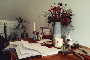 A horizontal colour photograph of a wooden table with a spread or notebooks, pens, and other random objects like a vase with dried flowers, candles, mugs, etc.