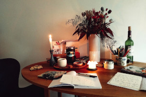 A horizontal colour photograph of a wooden table with a spread or notebooks, pens, and other random objects like a vase with dried flowers, candles, a tea tray with tea paraphenalia, etc.