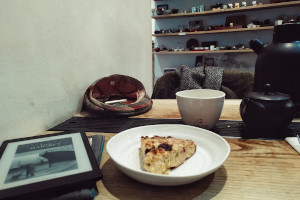 A horizontal colour photograph of a table at a tea shop with a slice of cake and an ereader at the front. Then there is a mug of tea and tea paraphernalia. In the background there are some shelves with various teaware on display.