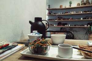 A horizontal colour photograph of a table at a tea shop with a white ceramic tray holding a salad in a small bowl and some gyoza cut off by the frame. There is a large mug of tea and tea paraphernalia behind it. In the background there are some shelves with various teaware on display.