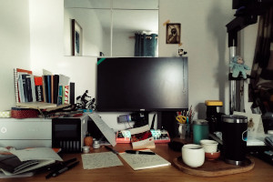 A horizontal colour photograph of a messy desk with papers, books, pens, and a tray with a tea set. Also on the desk, a printer, an enlarger, toys, and a screen and keyboard.