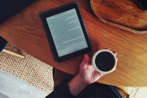 A horizontal colour photograph of a wooden table with an ereader on, a wooden tray, and a white hand on which a small cup of tea rests.