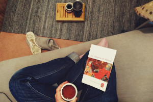 A horizontal colour photograph of crossed legs in blue jeans on a grey sofa. On one knee rests a book (Chanson de Roland). In the middle of the legs, a white hand holds a cup of tea. Below the sofa there is a salmon carpet mostly covered by a blue grey carpet. On it rests a bamboo tea tray with some tea paraphernalia.