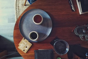 A horizontal colour photograph of a wooden table with a bluish plate at the edge on which rests some tea accoutrement. Around the plate is a wooden puzzle, a felt ereader cover, a large glass of water, and some pens and notebooks (those are cut off at the edge of the frame).