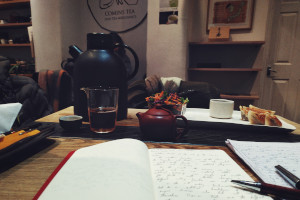 A horizontal colour photograph of a table with tea accoutrement, notebooks, pens, and a tray of food in the background.