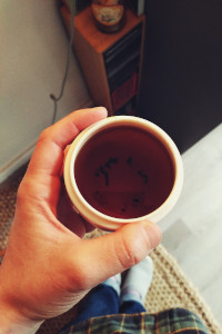 A vertical colour photograph of a cup of dark tea held in the hand over a half carpeted floor, half hard floor. In the background there is a small CD tower.